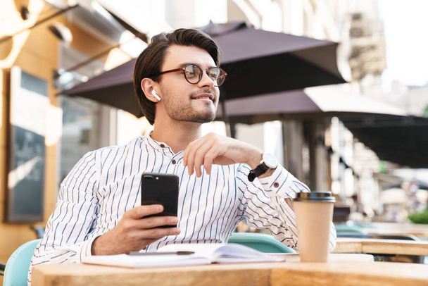 Photo of smiling caucasian man wearing eyeglasses using earpod and cellphone while working in cafe outdoors - Foto, afbeelding