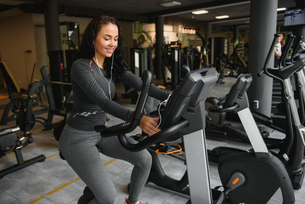 Young woman with headphones doing exercises on stationery bicycle in a gym or fitness center. Young sporty woman in gym listen music from smartphone. Women doing cardio exercises. - Photo, Image
