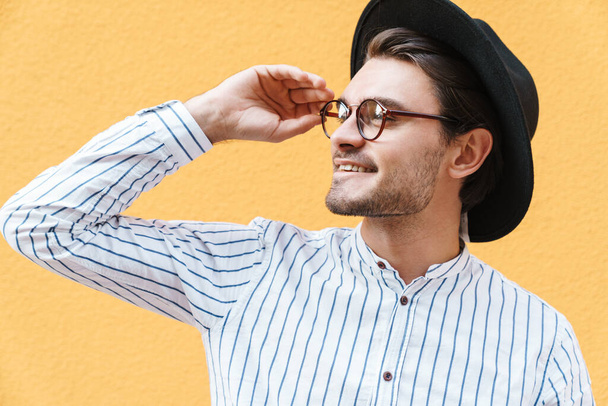 Image of young happy man wearing eyeglasses and black hat smiling and looking aside at copyspace isolated over yellow background - Foto, Imagem