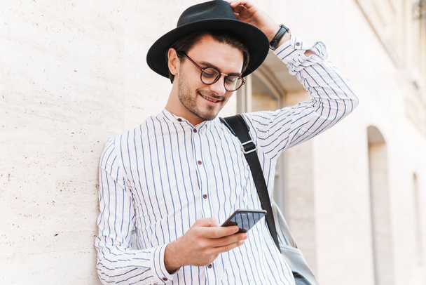Photo of cheerful caucasian man wearing eyeglasses and black hat typing on cellphone and smiling while leaning on wall in city street - Zdjęcie, obraz