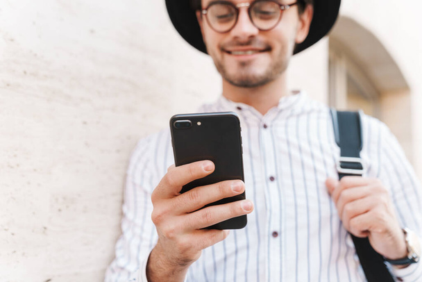 Photo of joyful caucasian man wearing eyeglasses and black hat typing on cellphone and smiling while leaning on wall in city street - Foto, imagen