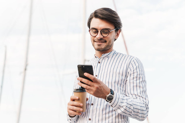 Photo of smiling young man wearing eyeglasses typing on cellphone and drinking coffee from paper cup while walking on pier - Photo, Image