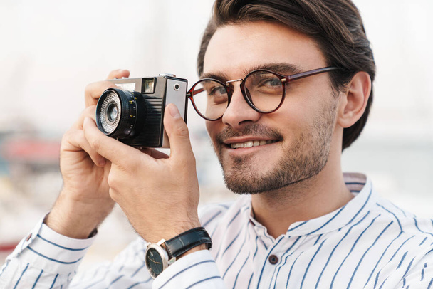 Photo of happy caucasian man wearing eyeglasses taking photo on retro camera and smiling while walking on pier - 写真・画像