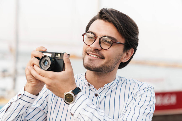 Photo of cheerful caucasian man wearing eyeglasses taking photo on retro camera and smiling while walking on pier - Zdjęcie, obraz