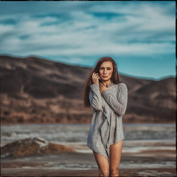 Young beautiful girl with dark hair in a gray sweater and sweater, stands among a deserted beach, a blue sky and cracked earth - Фото, изображение