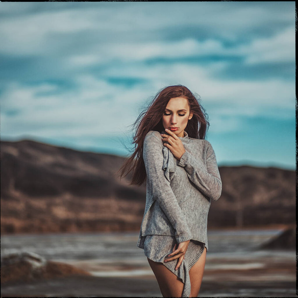 Young beautiful girl with dark hair in a gray sweater and sweater, stands among a deserted beach, a blue sky and cracked earth - Foto, imagen