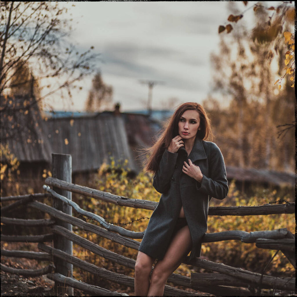 Young beautiful girl with dark hair in a black coat nude, stands near a wooden fence, against the background of autumn paints and an old house - Foto, Imagen