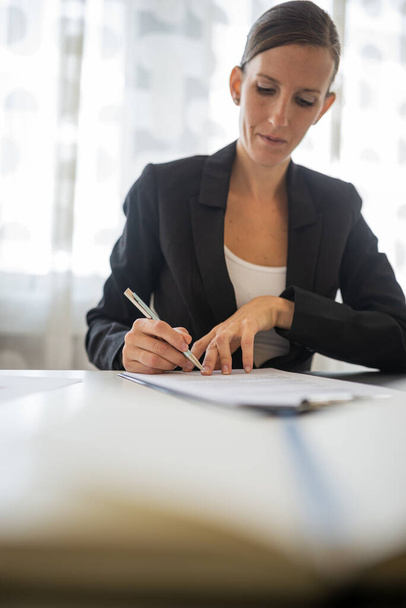 Poważna młoda bizneswoman siedząca przy biurku podpisująca dokument. - Zdjęcie, obraz
