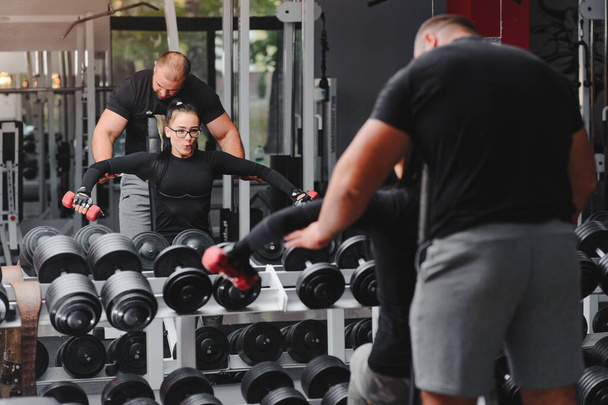 Fitness, sports, exercises and weightlifting. Concept - a young woman and a young man with dumbbells sweeping muscles in the gym. - Foto, afbeelding