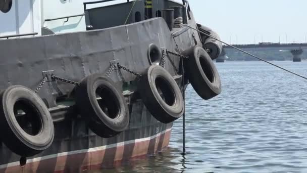Black side of port tugboat with car tires, anchored in port against background of large bridge over river - Footage, Video