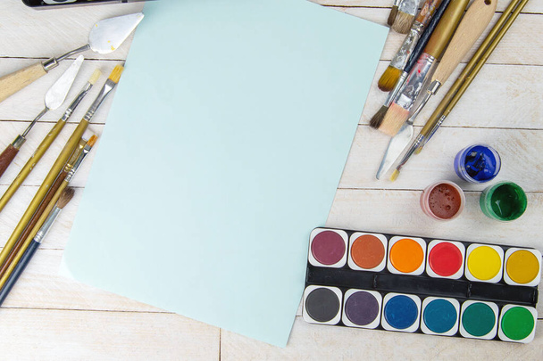 アーティストアクセサリーコレクションのセット。キャンバス、油絵具のチューブ、アートブラシ、パレットナイフと木製のテーブルの上に横たわる青い紙。アーティスト・ワークショップの背景. - 写真・画像