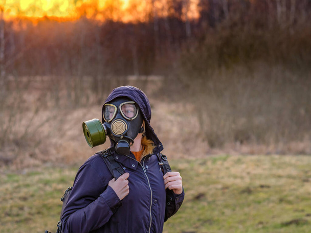 COVID-19. Girl in a black gas mask and dark clothes with a hood at sunset on a background of spring forest. On the back of the girl is a backpack. She flees a city infected with a viral infection.  - 写真・画像