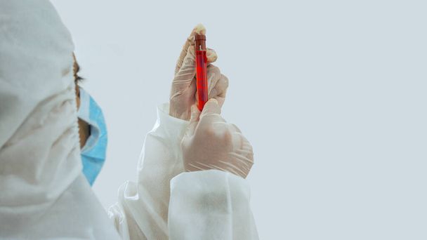 Vista posterior del médico con máscara y traje bioprotector sosteniendo un tubo de ensayo con etiqueta Covid-19 con muestra de sangre de coronavirus flotando dentro del tubo con la mano y mirando fijamente sobre fondo blanco
 - Foto, Imagen