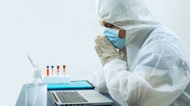 Doctor with blue mask and bioprotective suit working with his laptop in his office with his head resting on his hands with an attitude of concern, prayer or concentration with bottle of alcohol gel and test tubes at the desk on white background - Photo, Image