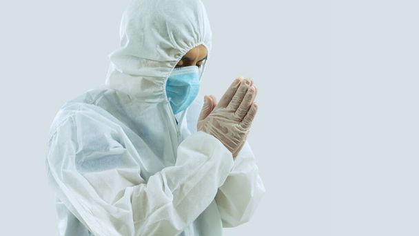 Zijaanzicht van arts met blauw masker en biobeschermend pak met hoofd naar beneden gekanteld en ogen dicht met handen samen met houding van bidden of mediteren op witte achtergrond - Foto, afbeelding