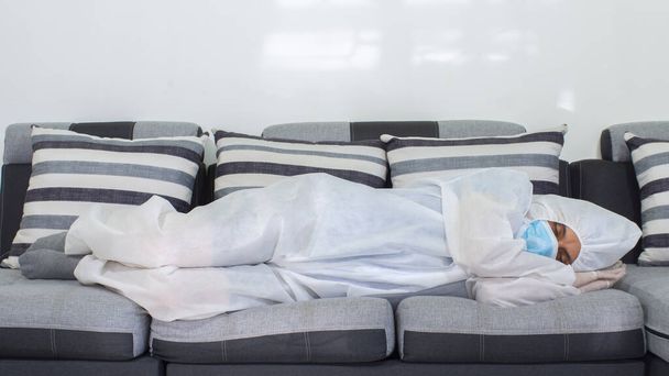 Előnézet orvos kék maszk és biológiailag védő ruha alszik egy nagy kanapén szürke tónusú éjszaka fehér háttér - Fotó, kép