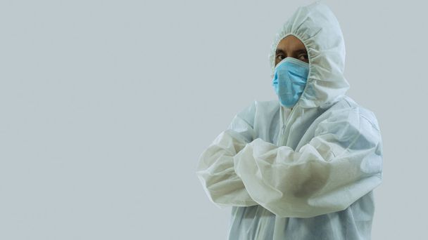 Vista lateral del médico con mascarilla azul y traje bioprotector blanco con brazos cruzados sobre fondo blanco
 - Foto, Imagen