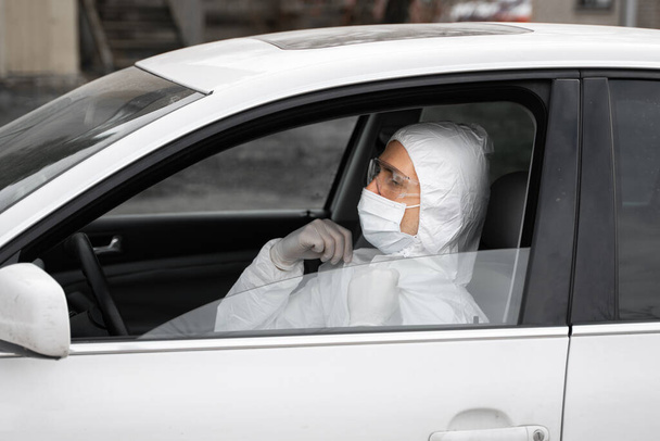 Человек в защитном костюме, медицинской маске и резиновых перчатках для защиты от бактерий и вирусов планирует водить машину. Защитная маска при карантине, мировая пандемия, ковид 19, коронавирус, инфекция
. - Фото, изображение