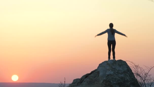 Silhouette di una donna escursionista che salta da sola su una grande pietra al tramonto in montagna. Turista femminile alzando le mani su alta roccia in natura sera. Turismo, viaggi e stile di vita sano concetto. - Filmati, video