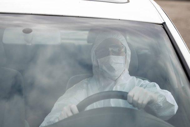 Man in protective suit, medical mask and rubber gloves for protect from bacteria and virus is driving a car. Protective mask while quarantine, world pandemic, covid 19, coronavirus, infection. - Foto, afbeelding