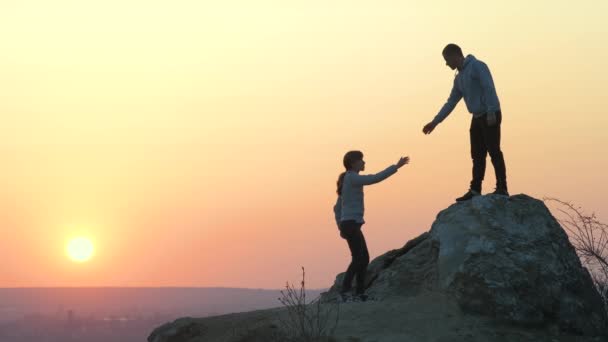 Man and woman hikers helping each other to climb stone at sunset in mountains. Couple climbing on high rock in evening nature. Tourism, traveling and healthy lifestyle concept. - Footage, Video
