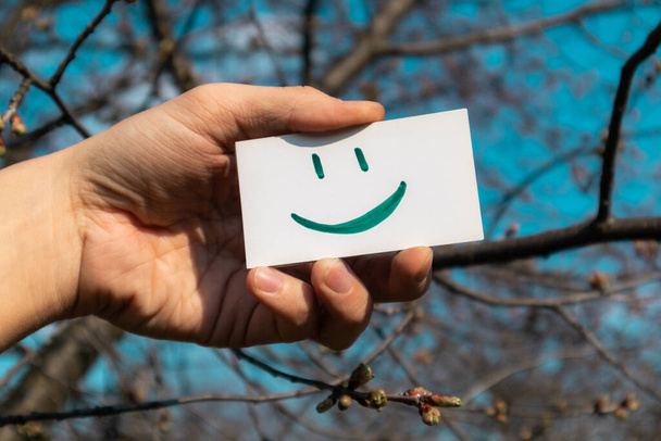 Boldog zöld mosoly emoji fehér papír kártya tartja a kezében a napos tavaszi égbolt színes fokozatú háttér. Pozitív hangulat jó idő - Fotó, kép