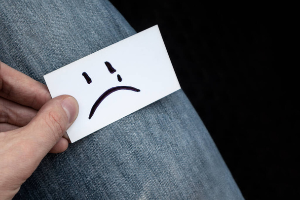 Emoji sorriso molto triste su carta bianca tenuta in mano su sfondo texture jeans. Triste stato d'animo cattivo tempo incolore
 - Foto, immagini