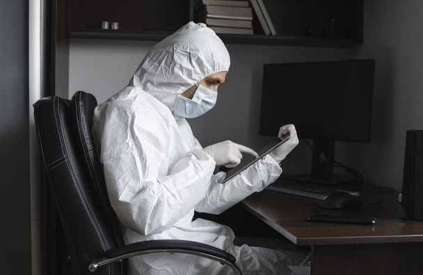 Man in protective suit, medical mask and rubber gloves sits at home and works with tablet at the table during quarantine. Designer, artist, architect, businessman at remote work in a pandemic covid. - Fotó, kép