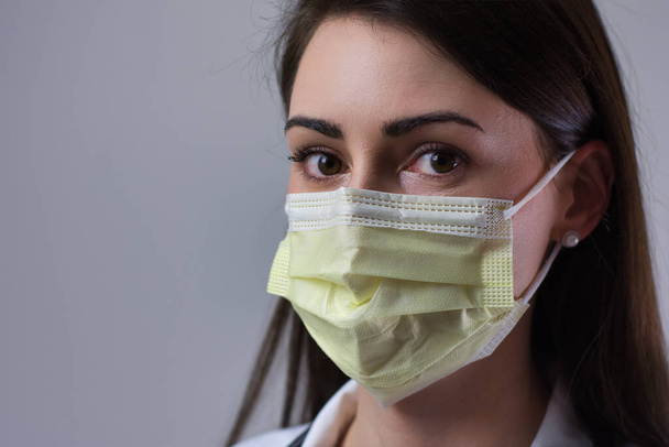 Female hospital worker wearing yellow face mask. Eyes visible over cover. Isolated on grey background. - Photo, Image