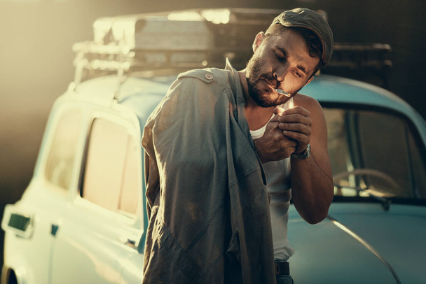 Mature bearded man lighting a cigarette, a retro car with suitcases on a roof rack on background - Φωτογραφία, εικόνα
