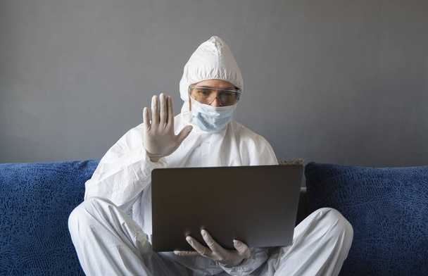 Man in protective suit, medical mask and rubber gloves sits at home and works with laptop at the table during quarantine. Designer, artist, architect, businessman at remote work in a pandemic covid. - Фото, изображение