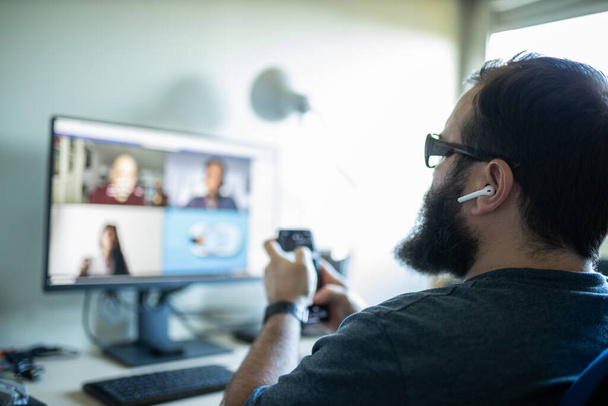 vousatý muž teleworking připojen videohovor se 3 lidmi a pomocí chytrého telefonu počítače a sluchátek - Fotografie, Obrázek
