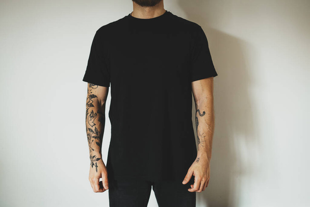 young attractive male hipster with a beard and tattoos, dressed in a black blank t-shirt, posing on a white wall background. Empty space for you logo or design. - Foto, imagen