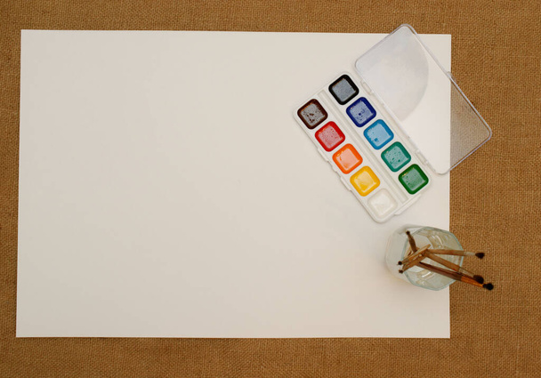New watercolor paints with brushes and a white sheet of paper. burlap brown background, Flat lay, space for text. - Foto, afbeelding