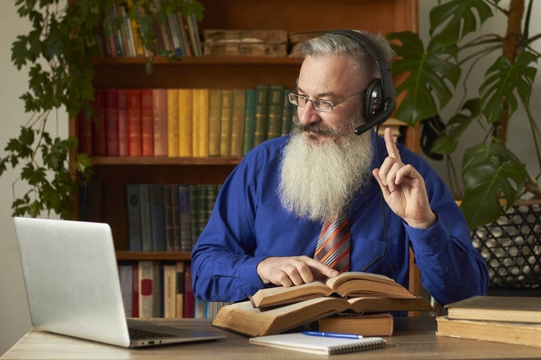 Distance Learning Concept. Teacher professor tutor in headphones teaches discipline online. Mature grey bearded man answers teacher's question through laptop. - Photo, Image