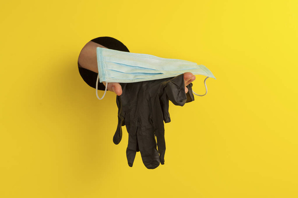 Disposable medical face mask and black gloves on the palm through a hole in a yellow background. Place for text. - Fotoğraf, Görsel