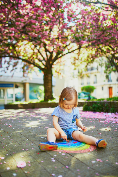 Adorable toddler girl drawing rainbow with colorful chalks on asphalt in park with blooming cherry blossom trees. Outdoor activity and creative games for small kids - Photo, Image