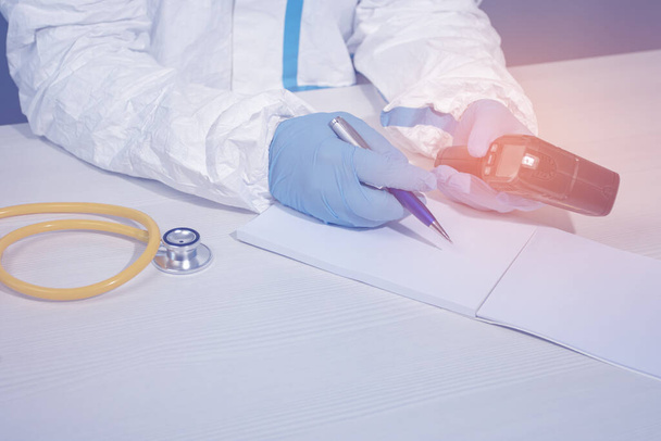 The doctor in a protective biological suit and gloves holds an infrared thermometer and a pen in his hand - Photo, Image