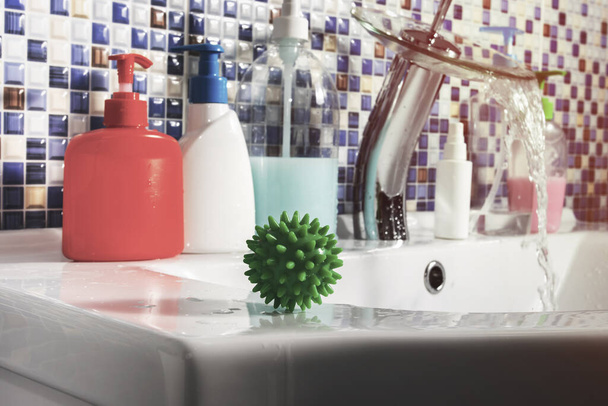 The green covid-19 coronavirus molecule sits on the white washstand in the bathroom. Lots of liquid soap in jars on the washstand. Against the background of running water from the tap - Foto, Imagen