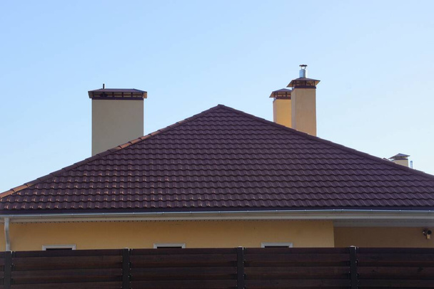 Красная черепица крыша частного дома с бетонными трубами за коричневым забором против голубого неба
 - Фото, изображение