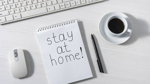 Op een wit bureau is een notitieblok met de woorden "thuis blijven" naast een toetsenbord, muis en een witte kop koffie, bovenaanzicht - Foto, afbeelding