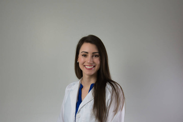 Woman healthcare professional in white coat isolated on grey wall. Head and shoulders visible. Attractive young lady with beautiful smile on neutral backdrop - Photo, Image