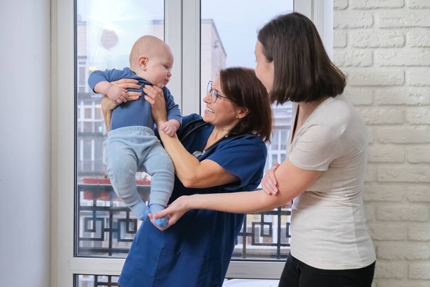幼児の子供を調べる女性医師。小児科医は彼女の母親と話をし、一緒に笑って、腕の中で赤ちゃんを取った。小児科、 1年までの子供のケアと健康 - 写真・画像