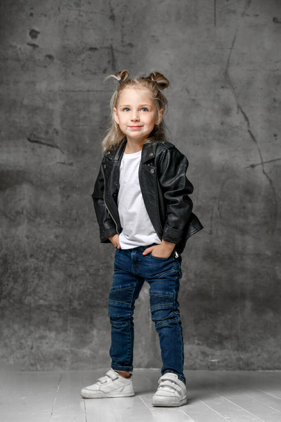Little smiling blond girl in stylish rock style black leather jacket, blue jeans and white sneakers standing with hands in pockets - Foto, Imagen