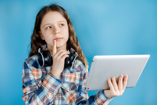 Teenagermädchen mit Kopfhörern beim Online-Schulunterricht zu Hause. Auf blauem Hintergrund. Mit nachdenklichem Gesichtsausdruck. Home Lernkonzepte - Foto, Bild