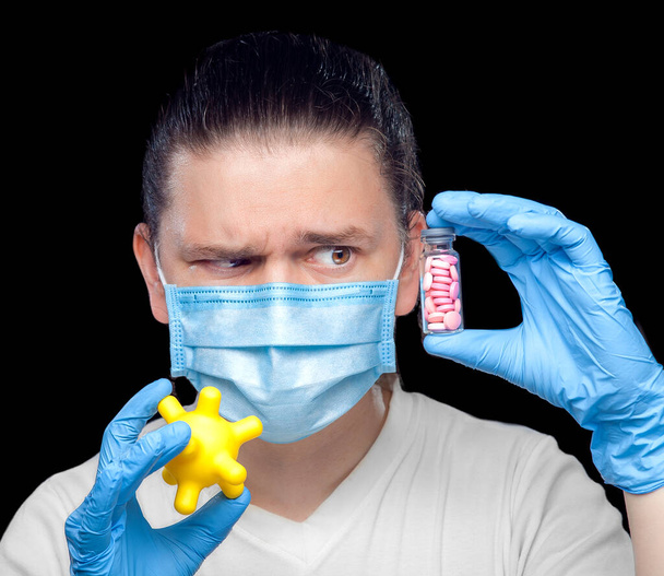 virologist in a medical mask and sterile gloves raised eyebrow contemptuously and looks at a glass bottle filled with pills for treating strain coronavirus, in his other hand is the bacteria covid-19. - Photo, Image