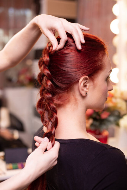 Beautiful, red-haired girl with long hair, hairdresser weaves a French braid, in a beauty salon. Professional hair care and creating hairstyles. - Photo, Image
