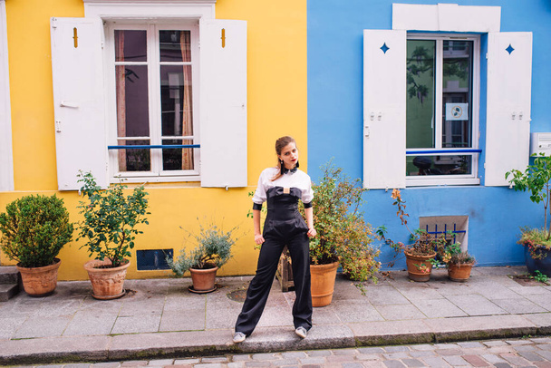 A beautiful young woman in a black fashionable jumpsuit stands on a Parisian street with colorful bright houses.France - Fotografie, Obrázek