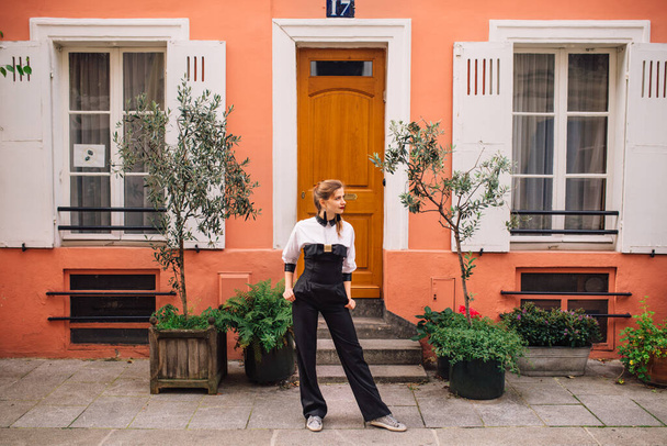 A beautiful young woman in a black fashionable jumpsuit stands on a Parisian street with colorful bright houses.France - Φωτογραφία, εικόνα