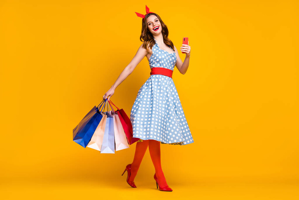 Full size photo cheerful girl shopping center client buy bargain hold bags go step use smartphone wear dotted blue skirt tights red headband high-heels isolated shine yellow color background - Photo, image